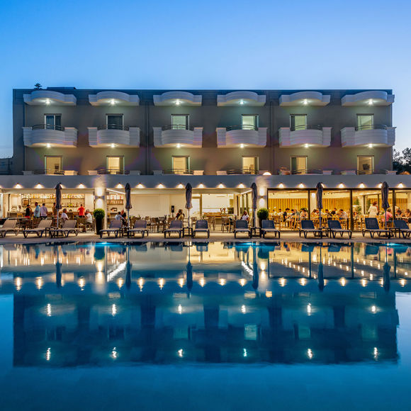Hotel exterior with illuminated balconies and restaurant area reflected in the pool at dusk