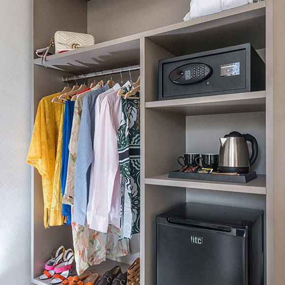 Open wardrobe featuring clothes, a safe, kettle, mugs, and a mini fridge