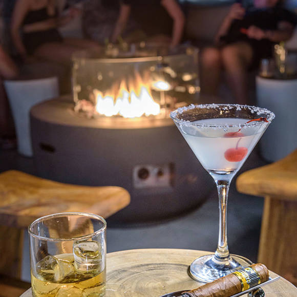 Close up of drinks and a cigar on a table with a firepit and people in the background