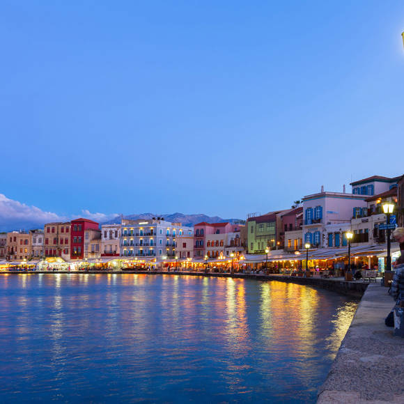 The waterfront road of Chania's Venetian harbor at dusk, lined with shops and restaurants